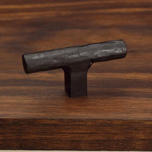 Peut inclure: Un bouton de meuble en forme de T avec une finition noire mate et une surface martelée texturée. Le bouton présente une barre cylindrique horizontale fixée sur une base carrée, offrant un aspect tactile distinct pour la quincaillerie.