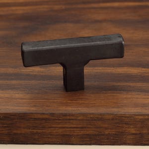 May include: A matte black T-shaped cabinet knob with a rectangular crossbar and a square base. The hardware features a solid, dark finish with clean, straight edges and a smooth, uniform surface texture, designed for attachment to a flat surface.