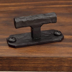 Peut inclure: Une poignée en forme de T en métal noir mat avec une finition martelée et texturée. La poignée présente une plaque de base rectangulaire fixée par deux vis cruciformes visibles, offrant une prise solide pour les portes ou tiroirs.