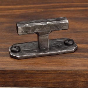 May include: A T-shaped cabinet knob with a dark grey, hammered metal finish. The knob features a horizontal bar attached to a rectangular base plate, which is secured to the surface by two black screws with visible cross-head indentations.