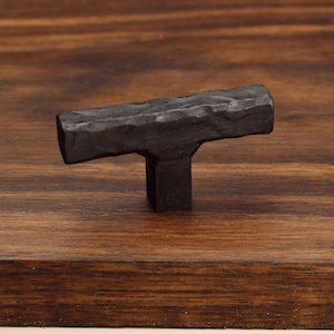 May include: A T-shaped cabinet knob with a dark, matte black finish and a textured, hammered surface. The hardware features a horizontal bar attached to a central square post, showcasing a rugged, uneven appearance across its entire metal body.