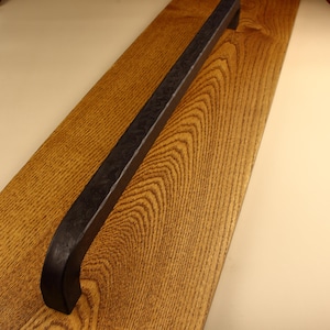 May include: A long, dark, matte black metal handle with a textured, hammered surface finish. The handle is mounted onto a rectangular piece of light brown wood that features a prominent, natural grain pattern running vertically along its length.