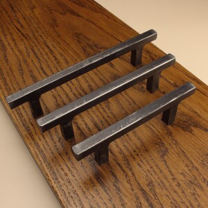 Peut inclure: Trois poignées de placard en métal gris foncé avec un design en barre rectangulaire et deux pieds de montage verticaux chacune. La quincaillerie présente une finition mate texturée et est disposée en rangée diagonale parallèle.