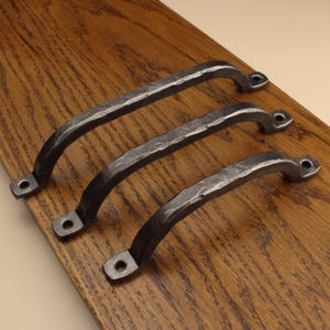 May include: Three dark grey metal drawer pulls with a hammered, textured finish. Each handle features a curved arch shape and two flat mounting ends, each containing a single circular screw hole for attachment to a surface.