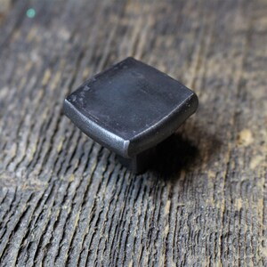 May include: A square, dark grey metal cabinet knob with rounded edges. The knob is set against a weathered, grey wooden surface. This hardware is suitable for drawers and cabinets.