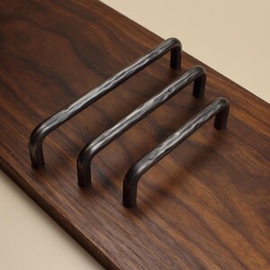 May include: Three dark grey metal cabinet pulls with a hammered texture. Each handle features a rounded U-shape design with two mounting points. The hardware is arranged in a diagonal row, showcasing the metallic finish and the dimpled surface.