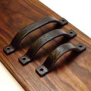 May include: Three dark grey, hand-forged metal drawer pulls with a textured, hammered finish. Each handle features a curved arch design with flat mounting ends, each containing a single circular screw hole for attachment to a surface.