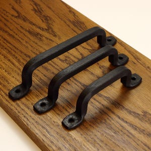 May include: Three matte black metal cabinet handles with a textured finish. Each handle features a curved grip and two flat mounting ends with pre-drilled holes for screws, arranged in a parallel row on a wooden surface for display purposes.