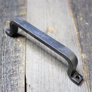 May include: A dark grey, forged iron handle with a curved design. The handle has a slightly textured surface and mounting holes at each end. It is shown on a weathered wooden surface. The handle is suitable for cabinets or drawers.