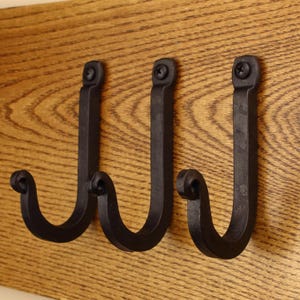 Peut inclure: Trois crochets en forme de J en métal noir avec des extrémités recourbées, chacun fixé sur une surface en bois clair avec une vis noire unique en haut. Les crochets présentent une finition mate et un design uniforme et robuste.