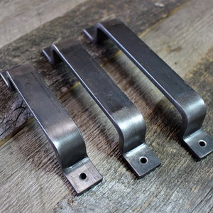 May include: Three dark metal door pulls with a curved handle and a rectangular base. Each handle has a single drilled hole for mounting. The door pulls are arranged on a weathered wooden surface. The handles are made of metal.