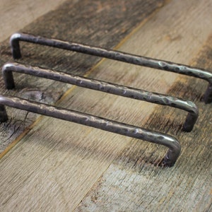 May include: Three hammered metal cabinet pulls with a dark, textured finish. The pulls are rectangular with rounded corners and a hammered surface. They are arranged on a weathered wooden surface.