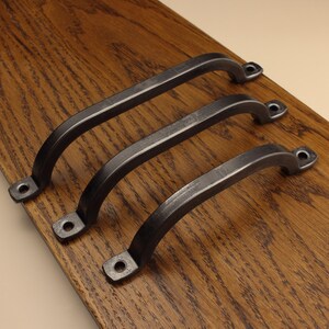 May include: Three dark grey metal cabinet handles with a curved, arched design. Each handle features two flat mounting ends with pre-drilled circular holes for installation, showcasing a smooth, solid construction with a consistent metallic finish.