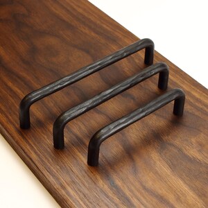 May include: Three dark charcoal-coloured metal cabinet pulls with a hammered, textured finish. Each handle features a rounded U-shape design with two mounting points, arranged in a diagonal row on a smooth, polished dark brown wooden surface.