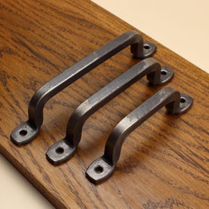 May include: Three dark grey metal cabinet handles with a textured finish. Each handle features a curved grip and two flat mounting ends, each containing a single circular screw hole for attachment to a surface. The metal has a matte appearance.
