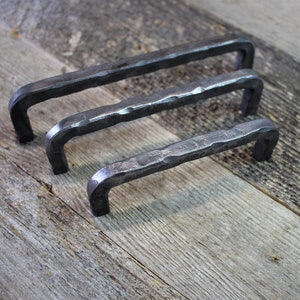 May include: Three black metal cabinet pulls with a hammered finish. The pulls are different sizes and are arranged in a row on a wooden surface.