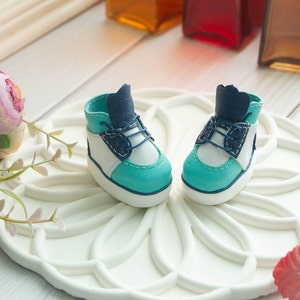 May include: A pair of miniature blue, white, and turquoise sneakers with white laces. The sneakers are sitting on a white decorative plate with a floral design.