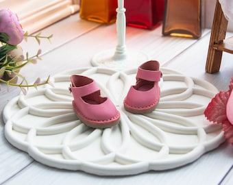 May include: A pair of pink leather doll shoes with a strap and buckle on a white decorative tray.