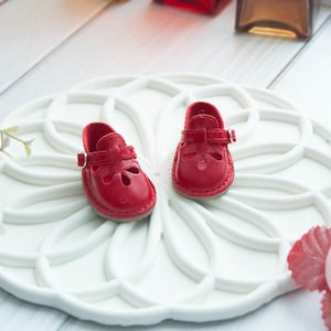 May include: A pair of red leather doll shoes with a buckle strap and cut-out detailing.
