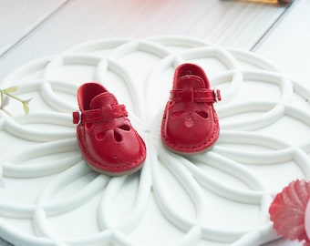 May include: A pair of miniature red shoes featuring a T-strap design with a small silver buckle and a flower-shaped cutout on the toe area. The shoes have a smooth finish and a light-colored sole, designed for a doll or small figurine display.