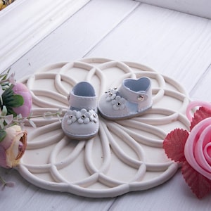 Puede incluir: Un par de zapatos pequeños de color blanco con una correa de cierre a presión metálico y una fila decorativa de flores recortadas en la parte delantera. Cada flor tiene un pequeño remache central plateado y una puntera redondeada.