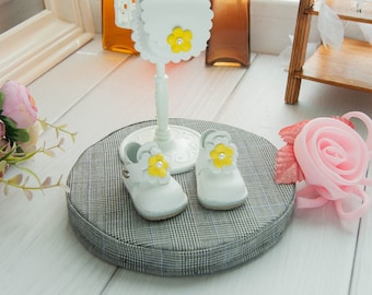 May include: A pair of white doll shoes featuring a yellow flower embellishment with a central white pearl accent on the toe. The shoes are displayed on a circular gray plaid cushion alongside a small white doll purse with a matching flower.