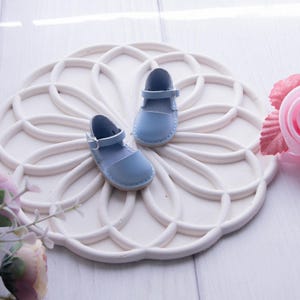 May include: A pair of light blue leather sandals with a strap and buckle closure. The sandals are on a white circular background with a decorative design.