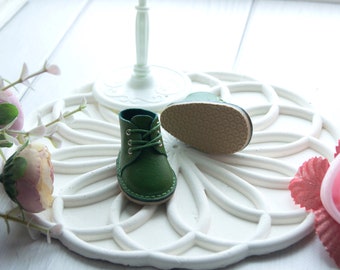 May include: A pair of miniature dark green leather boots with matching green laces and silver-toned eyelets. One boot is standing upright, while the other is positioned on its side to reveal a textured, tan-colored rubber sole with a circular pattern.