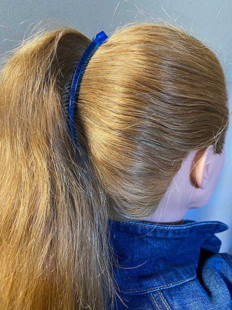 Puede incluir: Un peine de pl&aacute;stico azul asegura una cola de caballo rubia. El peine es arqueado y tiene una serie de dientes. El cabello est&aacute; peinado en una cola de caballo alta, y el modelo lleva una chaqueta vaquera azul.