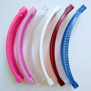 Puede incluir: Seis pinzas de pelo tipo banana curvadas en varios colores: rosa intenso, rosa claro, transparente, blanco, rojo y azul. Cada pinza tiene una serie de dientes a lo largo de la curva interior y un cierre en cada extremo. El texto "PAT 3998233" est&aacute; impreso en cada pinza.