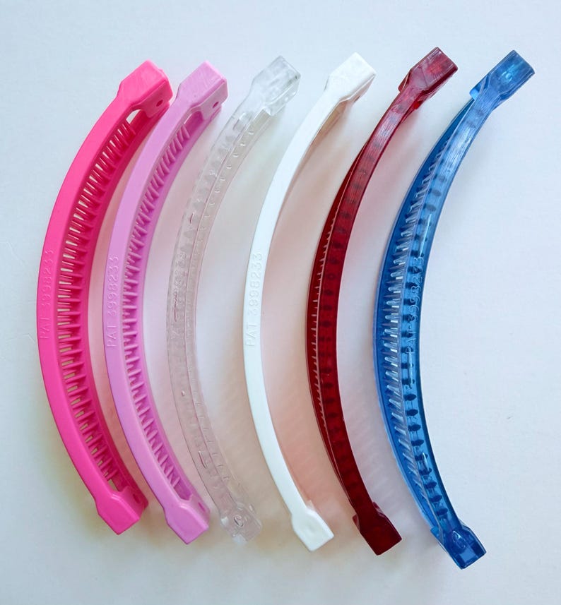 Puede incluir: Seis pinzas de pelo tipo banana curvadas en varios colores: rosa intenso, rosa claro, transparente, blanco, rojo y azul. Cada pinza tiene una serie de dientes a lo largo de la curva interior y un cierre en cada extremo. El texto "PAT 3998233" est&aacute; impreso en cada pinza.