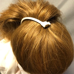Puede incluir: Peinado de cola de caballo con cabello casta&ntilde;o claro asegurado con una goma blanca. La goma para el pelo tiene un peque&ntilde;o elemento decorativo. El cabello est&aacute; peinado en un mo&ntilde;o, con la goma visible.