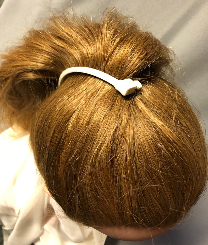 Puede incluir: Peinado de cola de caballo con cabello casta&ntilde;o claro asegurado con una goma blanca. La goma para el pelo tiene un peque&ntilde;o elemento decorativo. El cabello est&aacute; peinado en un mo&ntilde;o, con la goma visible.