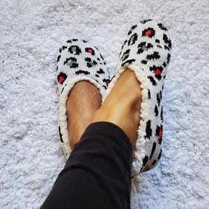 May include: A pair of knit slippers featuring a white base with a black and red leopard print pattern. The slippers have a soft, cream-colored fleece lining visible along the interior edges and a rounded toe design for a comfortable fit.