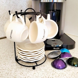 May include: A black metal espresso cup holder with white ceramic cups that say "Espresso" on them. There are also several colorful espresso pods on the counter.