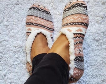 May include: A pair of knit slipper socks featuring a multi-colored fair isle pattern in shades of peach, grey, white, and black. The slippers are lined with a thick, white, plush faux-fur material and have a rounded toe design.