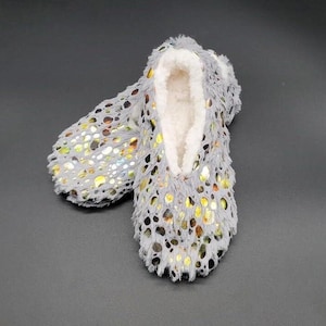 May include: A pair of gray, fuzzy slipper-style house shoes with a white, plush interior. The exterior features a pattern of gold and silver metallic accents. The slippers are designed for indoor wear and provide comfort and warmth.
