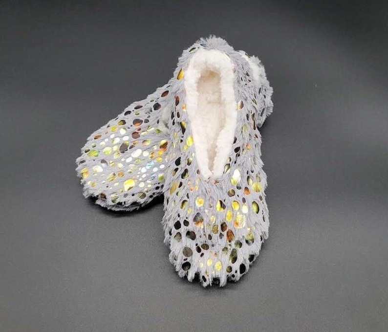 May include: A pair of gray, fuzzy slipper-style house shoes with a white, plush interior. The exterior features a pattern of gold and silver metallic accents. The slippers are designed for indoor wear and provide comfort and warmth.