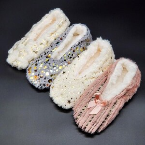May include: Four pairs of cozy slipper socks in cream, gray, white, and pink. Each slipper features a soft, plush interior and is adorned with decorative elements like gold stars and a bow. These slippers are designed for indoor comfort.