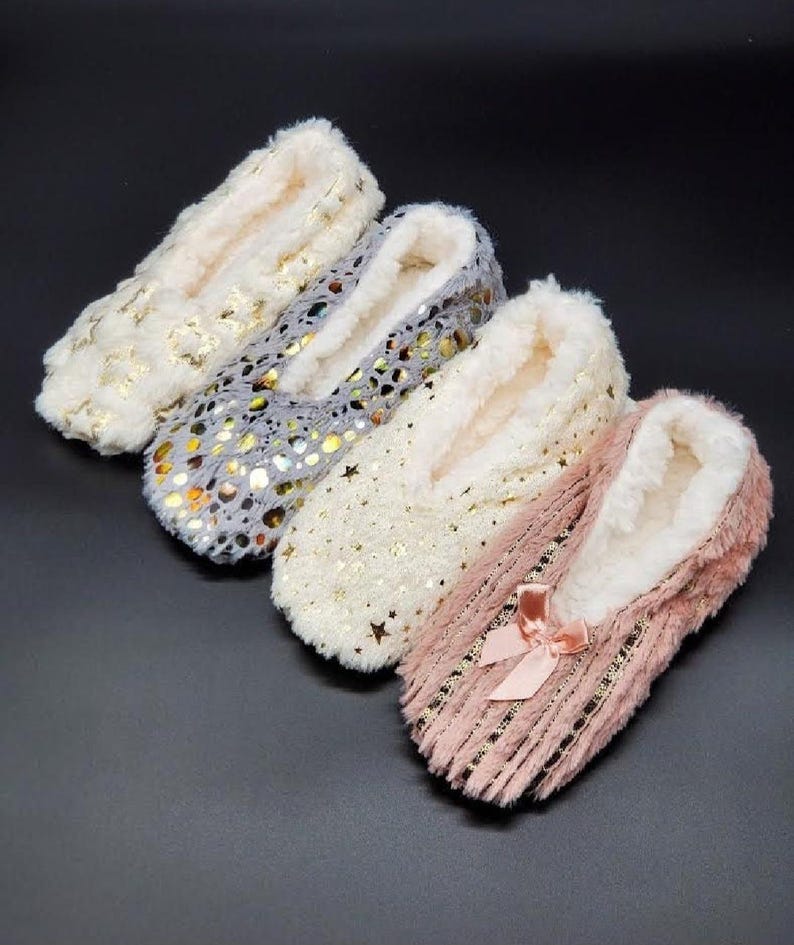 May include: Four pairs of cozy slipper socks in cream, gray, white, and pink. Each slipper features a soft, plush interior and is adorned with decorative elements like gold stars and a bow. These slippers are designed for indoor comfort.