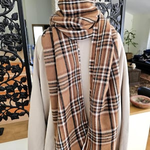 May include: A brown and white plaid scarf with black accents is draped around a mannequin's neck. The scarf has a fringed edge and is made of a soft, woven material. The scarf is displayed on a beige sweater.