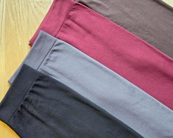 May include: A set of four pairs of leggings featuring a wide, ribbed elastic waistband. The leggings are presented in a stacked arrangement and come in solid colors including dark brown, deep burgundy, slate gray, and solid black.