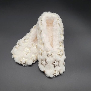 May include: A pair of ivory-colored, plush slippers with a gold star pattern. The slippers have a soft, fuzzy texture and are designed for indoor wear. The star pattern is evenly distributed across the surface.