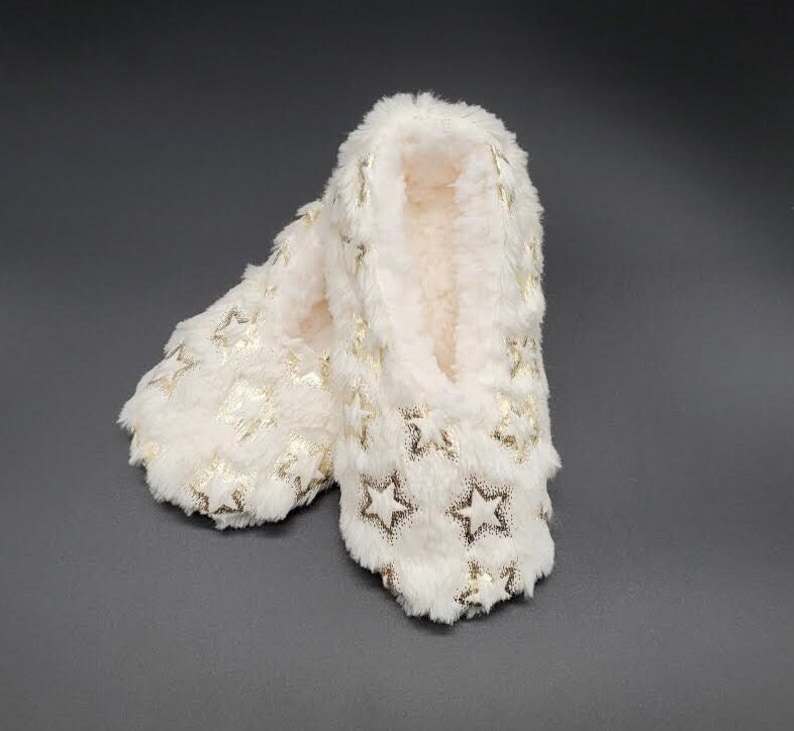 May include: A pair of ivory-colored, plush slippers with a gold star pattern. The slippers have a soft, fuzzy texture and are designed for indoor wear. The star pattern is evenly distributed across the surface.
