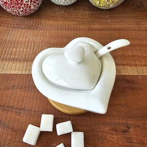May include: White ceramic heart-shaped sugar bowl with a lid and a small spoon. The bowl is sitting on a wooden surface with sugar cubes scattered around it.