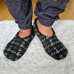 May include: A pair of black and gray patterned slippers with a soft, fuzzy texture. The slippers are worn on a white fluffy rug.