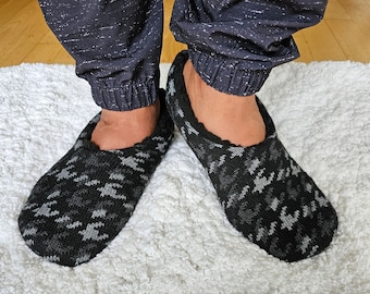 May include: A pair of black and gray patterned slippers with a soft, fuzzy texture. The slippers are worn on a white fluffy rug.