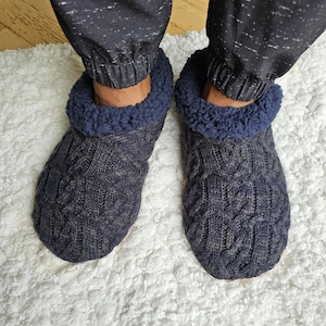 May include: A pair of dark charcoal grey cable knit slippers featuring a thick, navy blue textured fleece cuff around the ankle opening. The slippers have a classic braided pattern across the top and are designed to provide full foot coverage.
