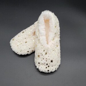 May include: A pair of ivory-colored slipper socks with a soft, plush exterior and a warm, fuzzy interior. The socks are adorned with gold star-shaped sequins, adding a touch of sparkle. These cozy slipper socks are designed for indoor wear.