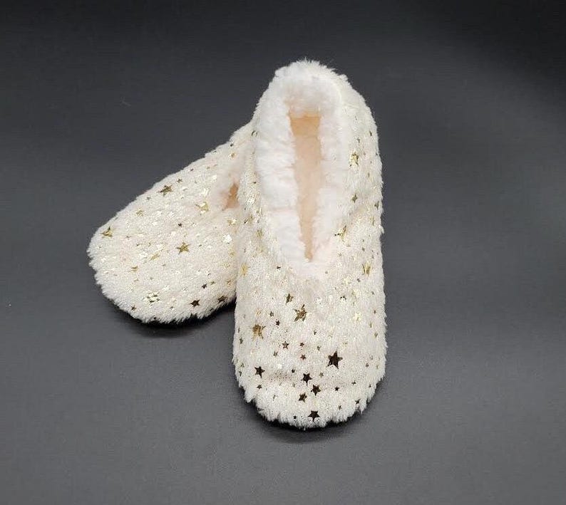 May include: A pair of ivory-colored slipper socks with a soft, plush exterior and a warm, fuzzy interior. The socks are adorned with gold star-shaped sequins, adding a touch of sparkle. These cozy slipper socks are designed for indoor wear.